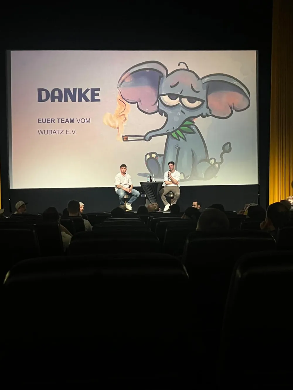 Zwei Vorstandsmitglieder von Wubatz e.V. sitzen auf der Bühne eines Kinosaals vor einer Leinwand mit der Aufschrift „Danke — euer Team vom Wubatz e.V.“ und dem Vereinsmaskottchen; davor die Sitzreihen des Publikums.